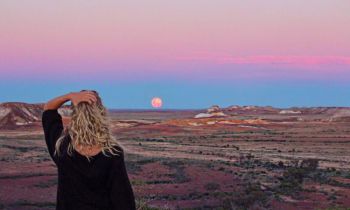 A girl with blonde hair in a black tshirt looking at sunset over an empty valley  in the Australian Outback on the 8 day Adelaide to Uluru adventure